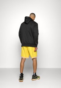 Felpa nera con cappuccio, pantaloni corti gialli con orlo beige, in piedi su uno sfondo neutro, che mette in evidenza il materiale e i colori dell'indumento.