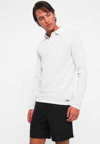 Young man wearing a white long-sleeve polo shirt and black shorts, looking to his left against a plain white background.