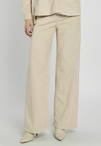 Beżowe spodnie w szerokiej nogawce z kordurowej tkaniny o prążkowanej teksturze i wysokim stanie, zestawione z pasującą koszulą z guzikami oraz spiczastymi botkami do kostek.