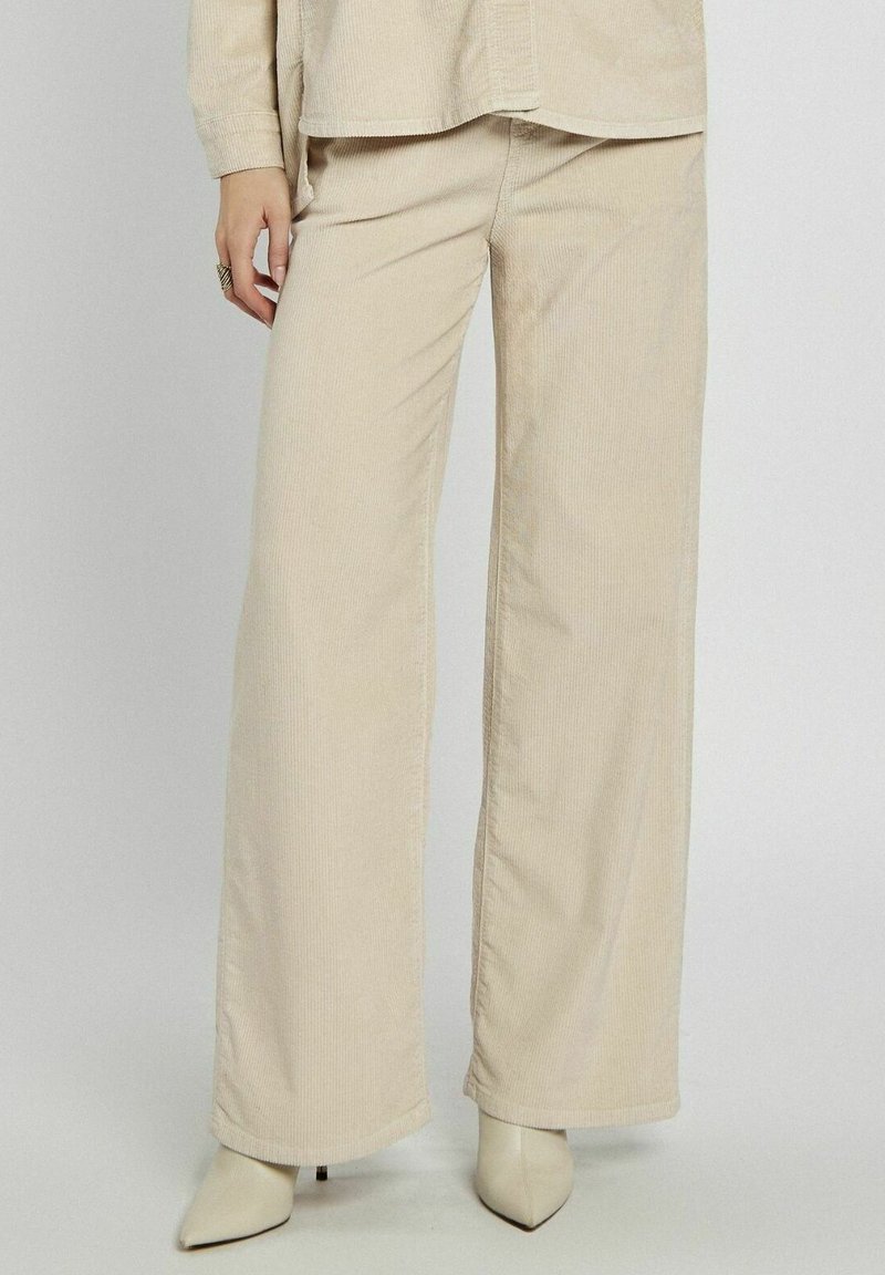 Beżowe spodnie w szerokiej nogawce z kordurowej tkaniny o prążkowanej teksturze i wysokim stanie, zestawione z pasującą koszulą z guzikami oraz spiczastymi botkami do kostek.