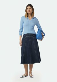 Himmelblauer, gerippter Pullover mit V-Ausschnitt, kombiniert mit einem dunkelblauen Jeansrock mit gelben Nähten. Das Model hält eine blaue Handtasche.