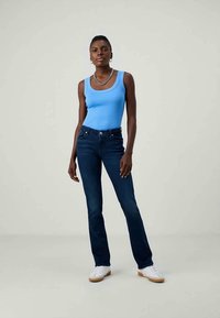 Mulher vestindo uma blusa sem mangas azul, jeans escuros, ténis brancos, colar de corrente prateado e brincos de argola, de pé contra um fundo simples.