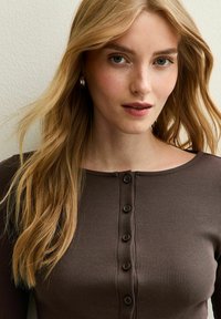 Bruine geribbelde top met lange mouwen, een ronde halslijn en knoopsluiting. De stof heeft een gestructureerde uitstraling, waardoor het aansluitende ontwerp extra wordt benadrukt.