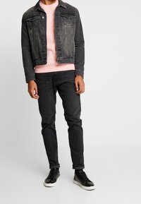 Mężczyzna w wyblakłej czarnej kurtce dżinsowej, różowej bluzie, czarnych dżinsach i czarnych trampkach z białymi podeszwami, stojący na tle jednolitego białego tła.