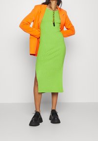 Abito midi a costine verdi con spacco laterale, abbinato a un blazer arancione brillante e scarpe chunk nere, con una collana di catena dorata.