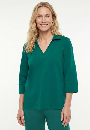 Blonde vrouw glimlacht, draagt een teal kleurige top met lange mouwen en een V-hals en bijpassende teal broek, staand tegen een witte achtergrond.