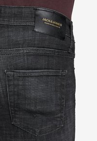 Jeans de mezclilla negra con una textura suave, que cuentan con un solo bolsillo trasero y un parche de marca de cuero con letras doradas.