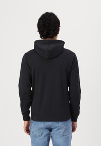 Uomo con capelli ricci che indossa un hoodie nero e jeans blu, in piedi di spalle contro uno sfondo chiaro e neutro.