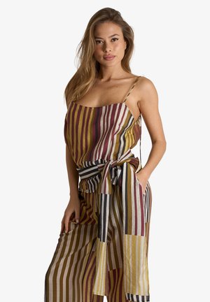 Giovane donna con capelli lunghi che indossa un vestito smanicato a righe multicolore legato in vita, in piedi con una mano in tasca.