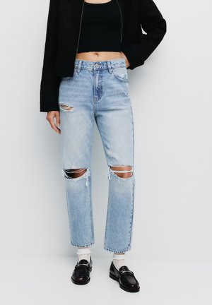 Lichtblauwe high-waisted jeans met verwerkte details, voorzien van grote kniebreuken en een rechte pijp. Gedeeltelijk gedragen met een zwarte jas en instappers.