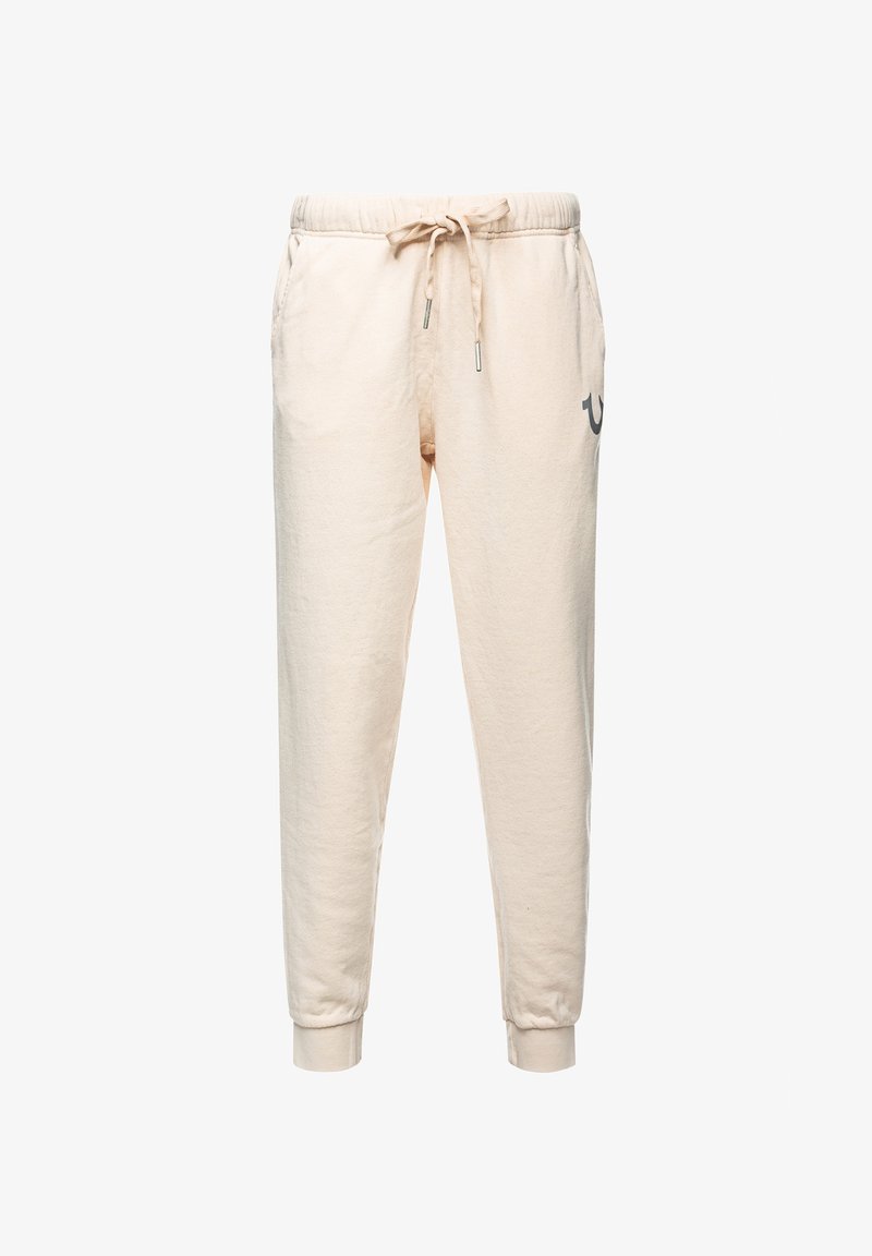 Beige joggingbukser lavet af blødt stof, med snøre i taljen, sidelommer og elastiske manchetter. Et logo er trykt i grå på benet.