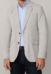 Homme portant un blazer gris clair sur une chemise bleu clair boutonnée et un jean bleu foncé, debout devant un fond uni.