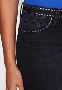 I jeans in denim neri presentano una cintura decorativa con borchie e hardware argentato. Il tessuto appare morbido con dettagli minimi sulle tasche.