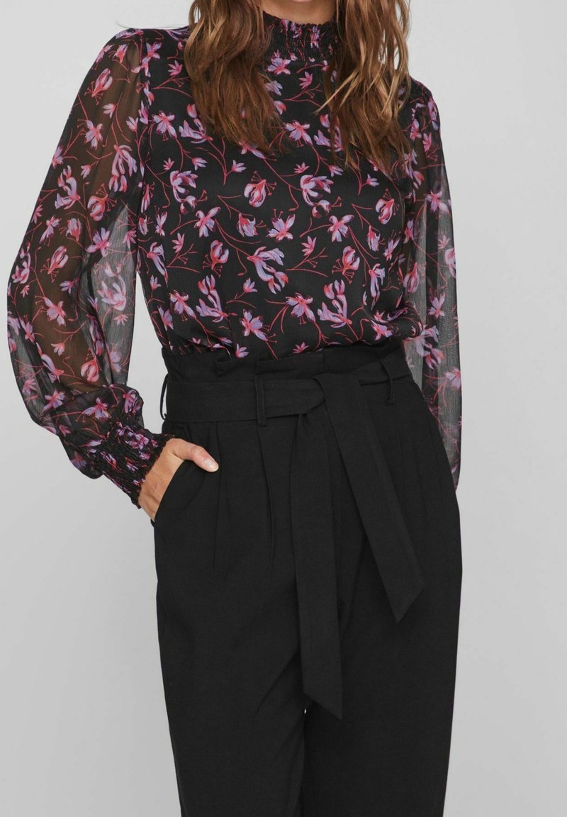 Femme portant un pantalon noir taille haute avec une ceinture nouée et un chemisier noir transparent à manches longues avec un motif floral rose.