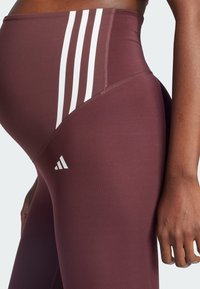 Bordowe legginsy z białymi trzema paskami wzdłuż biodra, o gładkiej fakturze i wysokim stanie. Logo Adidas na udzie.