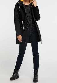 Svart vattentät parka med en gul huva på insidan, med dragsnören, två framfickor och en knälång design. Matchas med mörka jeans och svarta stövlar.