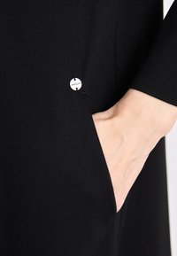 Svart plagg med en glatt tekstur, med en sidelomme og en sølvknapp med gravering. Designet fremhever en minimalistisk stil.