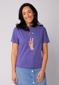 Lila Baumwoll-T-Shirt mit einem Grafikdesign einer Hand, die ein Friedenssymbol zeigt, und bunten Herzformen darüber. Kurze Ärmel und Rundhalsausschnitt.