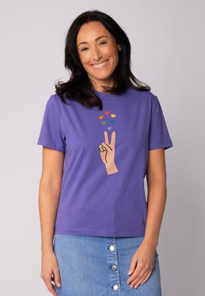 T-shirt en coton violet arborant un graphique d'une main faisant un signe de paix, avec des formes de cœurs colorées au-dessus. Manches courtes et col rond.