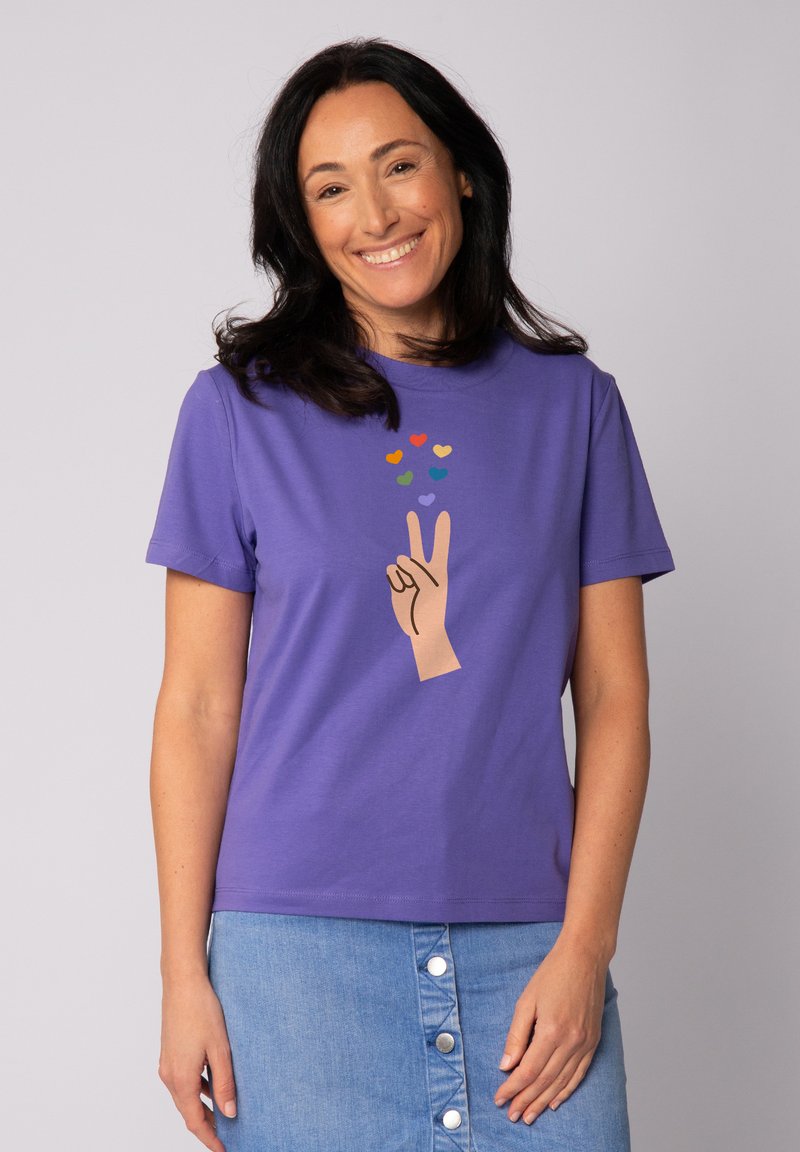 Lila Baumwoll-T-Shirt mit einem Grafikdesign einer Hand, die ein Friedenssymbol zeigt, und bunten Herzformen darüber. Kurze Ärmel und Rundhalsausschnitt.