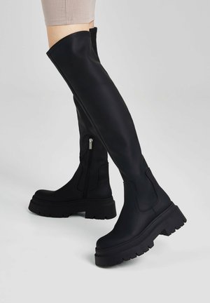 Bottes noires over-the-knee en matériau lisse, dotées d'une fermeture éclair sur le côté, d'une semelle épaisse et de coutures renforcées en haut et sur les côtés.