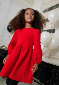 Sarkans kleita ar teksturētu audumu, ar balonu piedurknēm un vidukļa šuvi. Fons ar melnām un baltām baloniem un marmora fonu.