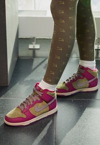 Tenisky Nike ve vysokém provedení mají design z růžového a olivově zeleného semiše, šedé akcenty a texturované podrážky, doplněné o vzorované legíny.