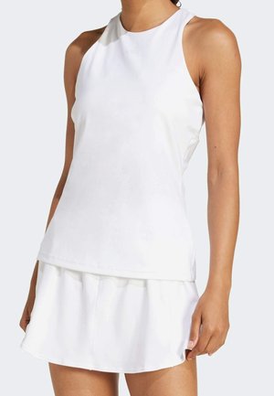 Femme portant un haut blanc sans manches et une jupe blanche assortie, debout devant un fond clair uni.