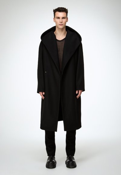 Langer schwarzer Wollmantel mit großer Kapuze, offenem Vorderteil und dezenten Knopfdetails. Getragen über einem braunen Hemd und kombiniert mit schwarzer Hose und Schuhen.