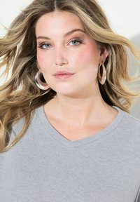 Grå v-hals sweater, glat tekstur. Model med bølget, skulderlangt hår, iført store sølvøreåbninger. Naturlig makeup, neutrale toner.
