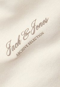 Logo Jack & Jones Archive Selection brodé sur un tissu beige texturé.