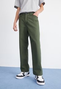 Green, straight-leg pants made of cotton, featuring a standard waistband, front pockets, and a smooth, matte texture. Paired with black and white sneakers.