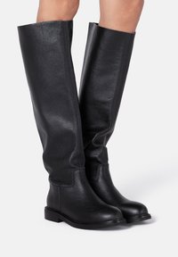 Bottes en cuir noir montant jusqu'au genou avec une finition texturée, bout arrondi et semelle plate noire. Tige lisse avec des détails de couture minimaux.