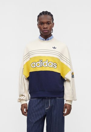 Młody mężczyzna noszący kolorową bluzę Adidas z granatowymi, żółtymi i kremowymi paskami oraz granatowe spodnie w cienkie paski na jednolitym tle.