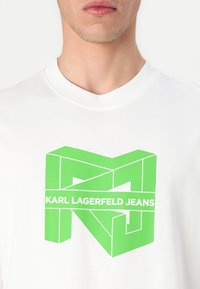 Bílá bavlněná tričko s výrazným zeleným geometrickým logem a textem "KARL LAGERFELD JEANS" umístěným uprostřed pod ním.