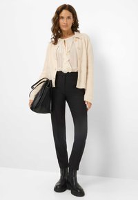 Cardigan a coste crema, blusa avorio arricciata, pantaloni neri su misura e stivaletti neri alla caviglia. La borsa ha una finitura opaca con lunghe cinghie.