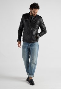 Zwarte leren jas met ritssluiting, twee voorkruiken en schouderflappen, gedragen over lichtblauwe spijkerbroek en zwarte schoenen.