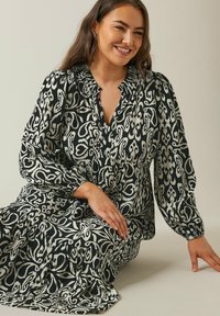 Schwarz-weiß gemustertes Kleid mit langen Ärmeln, einem gerafften Ausschnitt und lockerer Passform. Der Stoff hat eine fließende Textur und ein geschichtetes Design.