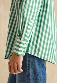 Grün-weiße gestreifte Button-Up-Bluse mit erleichtertem Schnitt, die markante Manschetten und eine glatte Baumwolltextur aufweist. getragen über blauen Denim.