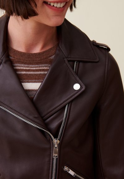 Veste de moto en cuir marron avec quincaillerie argentée, fermetures éclair diagonales et épaulettes. Superposée sur un pull en tricot marron à rayures.