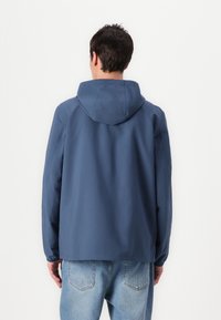 Jachetă bleu marin cu glugă, dintr-un material neted. Are o croială lejeră și mâneci lungi. Purtați-o peste blugi de culoare albastru deschis.