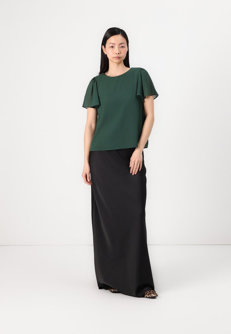 Blouse verte avec des manches courtes volantées, associée à une longuejupe maxi noire fluide. La blouse a un col rond et une texture douce.