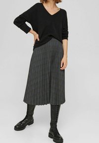Suéter negro de cuello en V, falda midi de cuadros grises con pliegues y botas negras hasta la rodilla con cordones, creando un look en capas y texturizado.