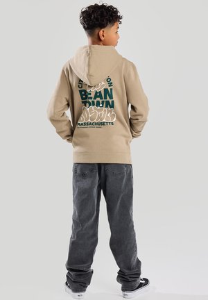 Beige Kapuzenpullover mit einem großen grünen Grafikdruck mit der Aufschrift "BOSTON BEANTOWN MASSACHUSETTS" auf dem Rücken, kombiniert mit lockeren grauen Hosen und schwarzen Sneakern.