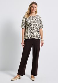 Krótkie rękawy, kremowa bluzka z czarnym abstrakcyjnym wzorem, zestawiona z brązowymi, szerokimi spodniami i beżowymi płaskimi butami. Gładka tekstura tkaniny.