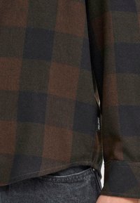 Camisa de franela a cuadros marrones y negros con mangas largas, aberturas laterales y dobladillos cosidos. Etiqueta visible cerca de la parte inferior en el lado izquierdo.