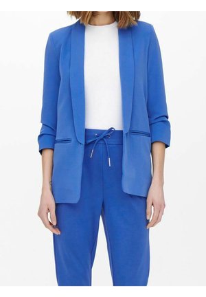 Vrouw draagt een felblauwe blazer met opgerolde mouwen over een wit shirt, gecombineerd met bijpassende blauwe broeken met trekkoord, getoond van nek tot heupen.