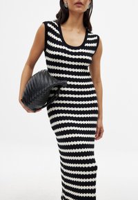 Robe tricotée rayée noir et blanc avec un décolleté en forme de scoop et une coupe ajustée. Le modèle tient une pochette tissée noire à texture.