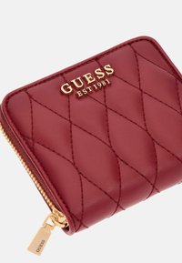 Rode gewatteerde leren portemonnee met een gouden ritssluiting en de merknaam "GUESS EST 1981" in goud op de voorkant gebosst. 