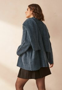 Cardigan épais, bleu foncé, avec des bordures côtelées, coupe oversize et deux poches à l'avant, superposé sur une jupe noire. Texture douce.